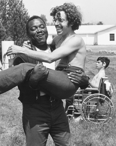 a man holds another in his arms, man behind them in a wheelchair. Field setting, shot in Black and white
