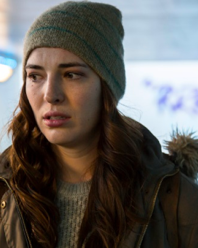 Young woman with brunette hair, beanie hat and fluffy hooded jacket stands somewhere which looks cold and grey. She looks scared and unsure of what may be behind her.