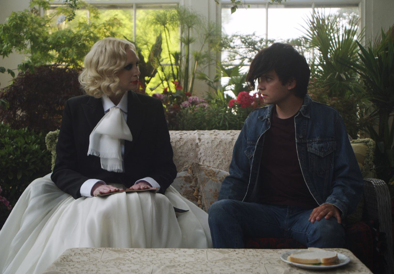 A woman and a young man face each other on a sofa in a room full of plants.