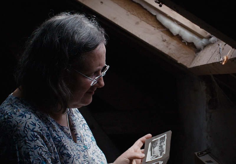 A woman stands in an attic and looks at an old photograph.