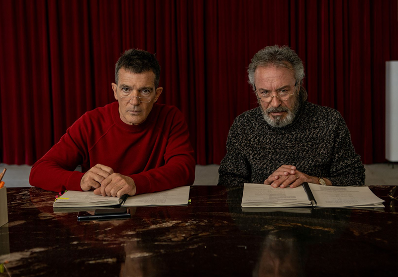 Two men sit at a table with script-books and look ahead.